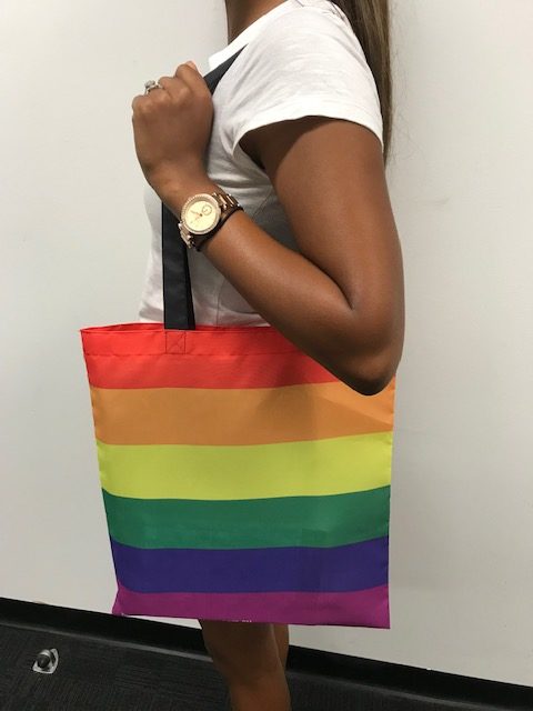 Image of tote bag in rainbow pride flag colours, hanging from shoulder of brown-skinned person.