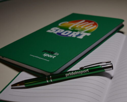 image shows a green and silver pen with the words Pride in Sport on the barrel, resting on the lined page of an open book, with another green notebook resting above it showing a green cover with the words Ally in Sport