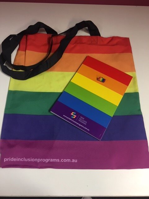 Image shows a rainbow coloured tote bag with black handles, with an A5 rainbow covered notebook, and a small rainbow lapel pin