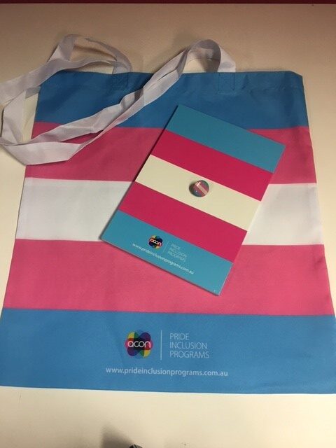 Image shows a tote bag in trans pride flag colours, with a matching A5 notebook and small lapel pin